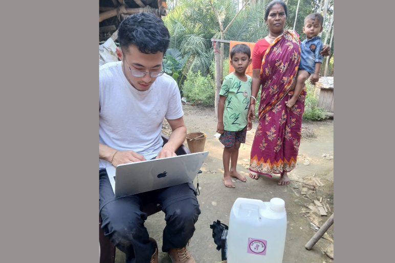Jon Bessettee conducts research on his laptop in a village in India with people from the village standing behind him.