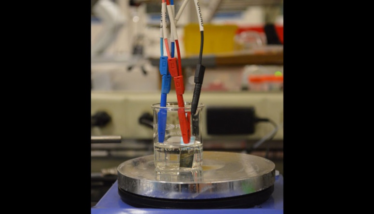 Blue, red, and black wires clamped to piece of metal in liquid in beaker