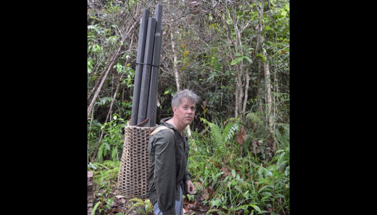Harvey carrying pipes on back through jungle