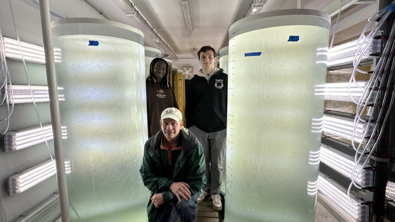 Three people, two standing, one crouching by two large algae tanks, over 6 feet tall in a room flanked with bright, fluorescent lighting on the sides of the walls