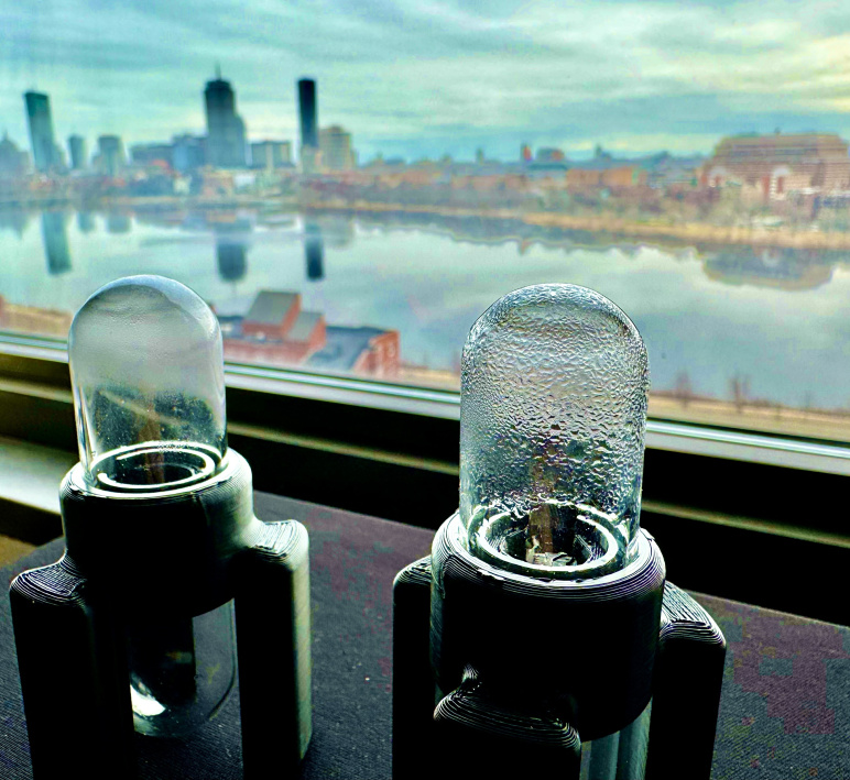 Two devices with glass enclosures, used for scientific measurements, next to a window with a view of Boston skyline.