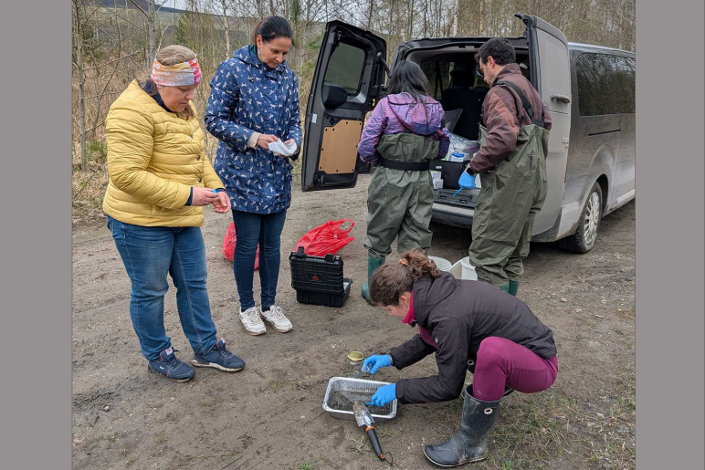 A group of people stand around the back of a van with double doors open, as one person crouches on the ground sifting soil samples in a pan