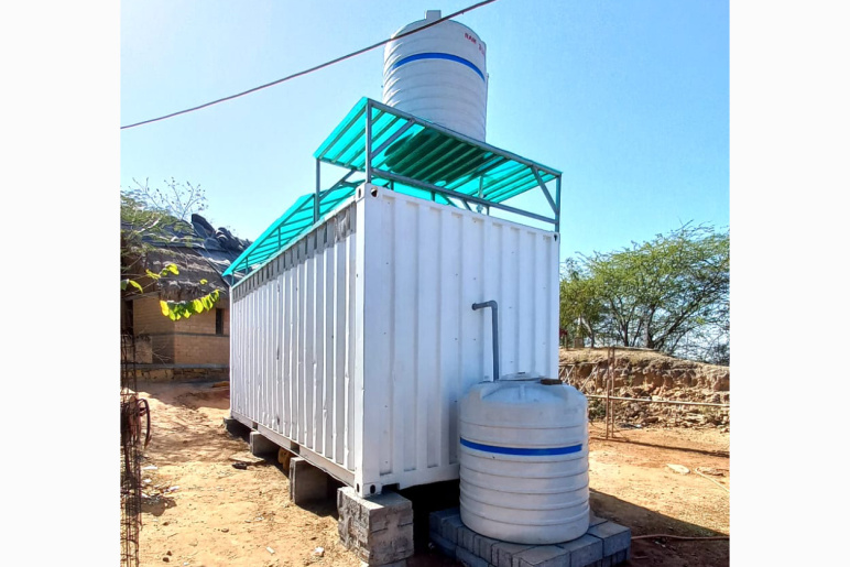 White, metal shipping container that has been fitted with a plastic roof and two water tanks, one on the roof and one on the ground behind the unit