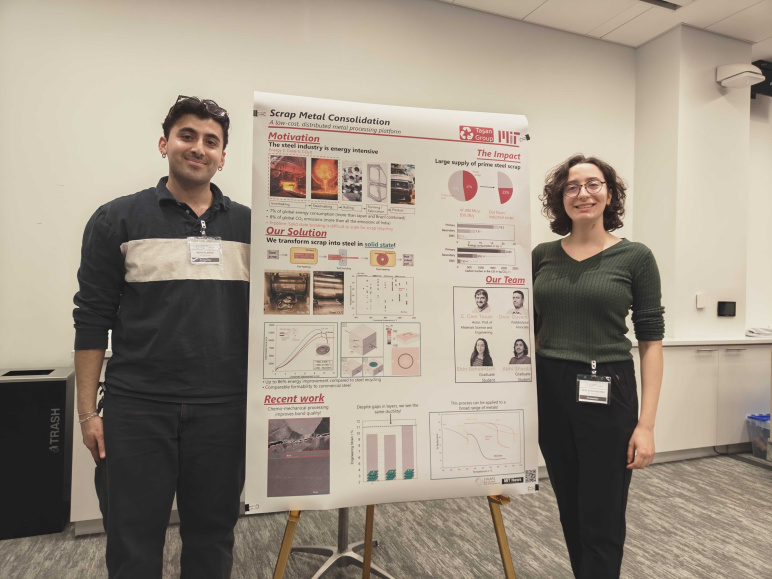 A male student stands to the left of a large poster on an easel and a woman stands on the right, both smiling