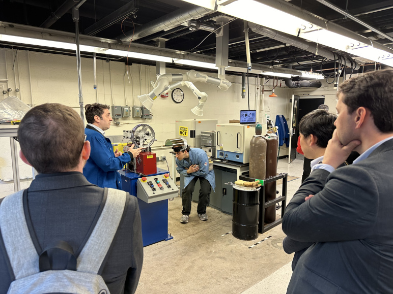 A group of people in a lab watch two researchers in blue lab coats conduct an experiment