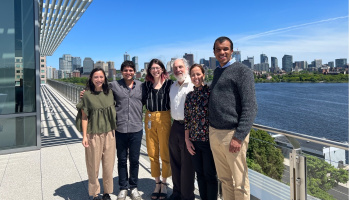 Group of six people standing on a roof deck outside, overlooking the Charles River and Boston