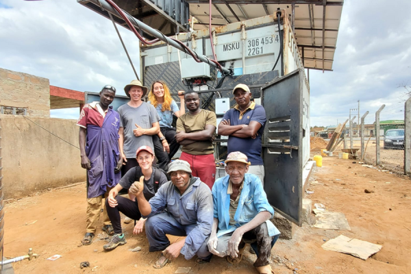 A group of 8 people, five standing and three crouching, posing in front of a shipping container being used for cold storage.
