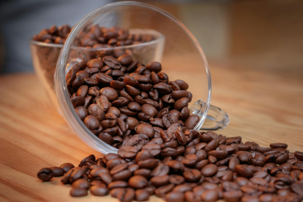 A clear coffee mug spilling out roasted coffee beans on a wooden table.