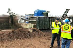 Two individuals wearing yellow vests standing in front of a composting facility