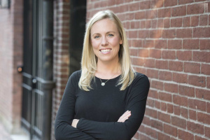 A woman with blonde hair wearing a black shirt crossing her arms and smiling in front of a brick wall. 