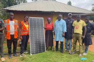 Individuals standing with a solar panel in front of a small building