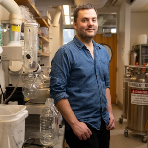 Daniel Suess stands in a lab with pipes and equipment behind him