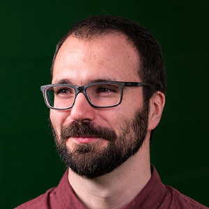 White man with dark beard, glasses and red shirt, looking to the side, off camera