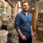 Daniel Suess stands in a lab with pipes and equipment behind him