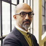Headshot of John E. Fernández, he wears glasses and stands near a light filled window
