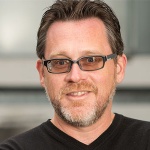 Man with pepper gray beard, brown hair, and glasses smiling