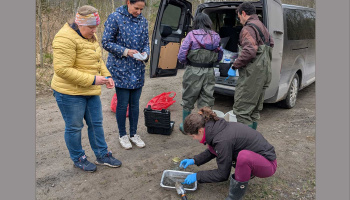 A group of people stand around the back of a van with double doors open, as one person crouches on the ground sifting soil samples in a pan