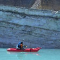 An Individual in a red kayak paddling on a turquoise lake near a towering ice wall with visible layers of sediment